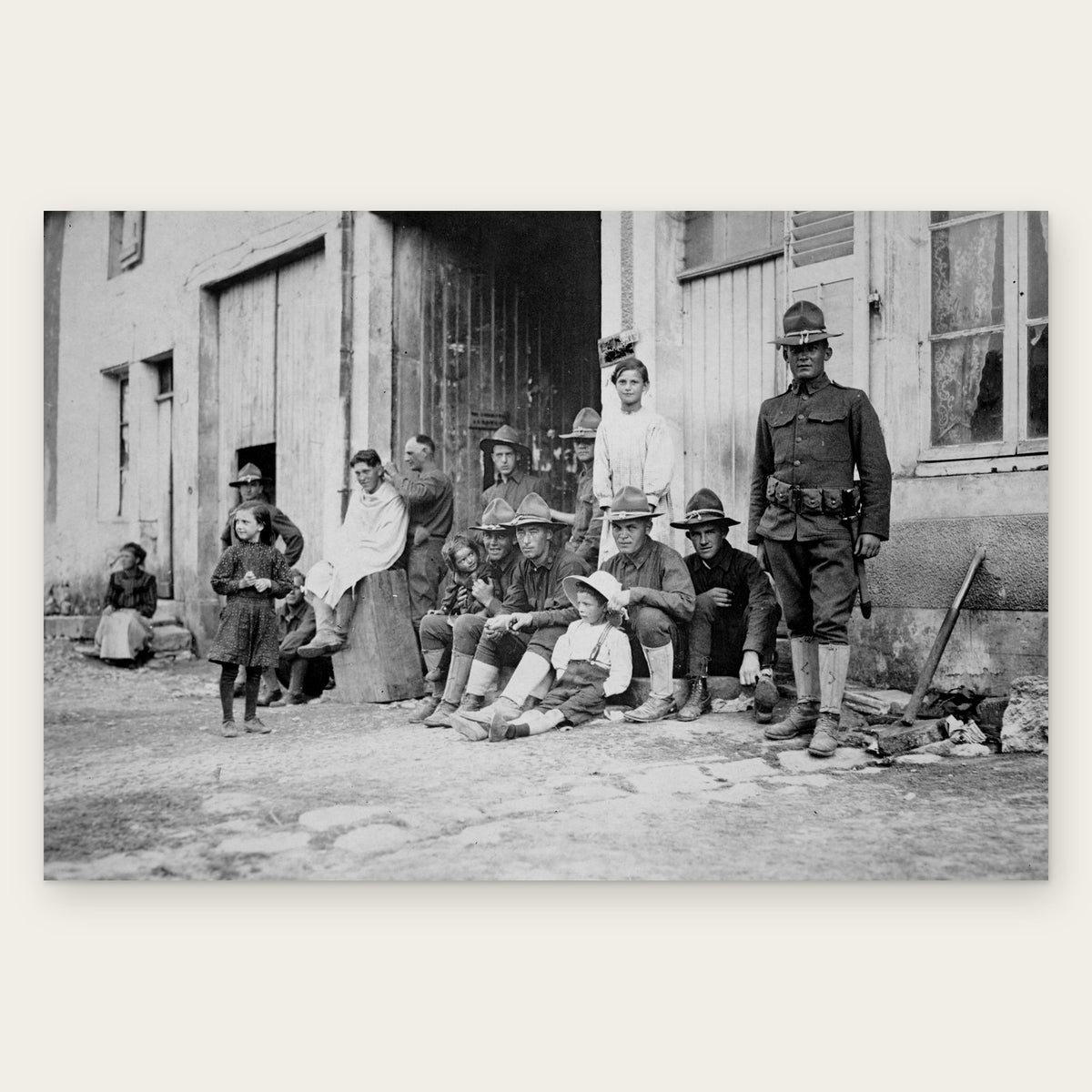 American soldiers in campaign hats sit with children outside a house in a French village while one soldier gets a haircut, restored vintage photo print