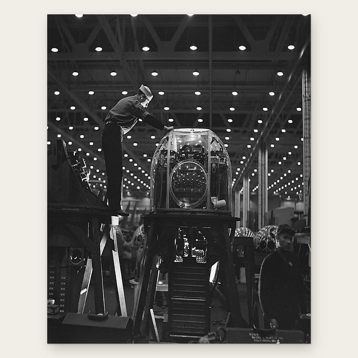 A worker in a white cap stands on a wooden platform and reaches across the top of a glass-domed machine gun turret on a test stand, under rows of factory lights, restored vintage photo print
