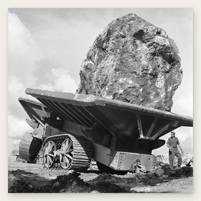 A huge rough boulder rides on a steel crawler wagon under a cloudy sky, with a Seabee in a cap standing beside the wagon's tracks, restored vintage photo print